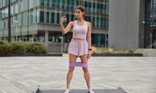 0 full length shot active slim woman records video her sport blog dressed tracksuit trains with resistance band poses fitness mat urban setting makes video call while having aerobics 1